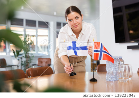 Young female assistant putting up norwegian and finnish flags on table for international negotiations 139225042