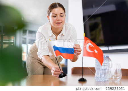 Positive young woman putting flag of Russia on the table with flag of Turkey in conference room Positive young woman putting flag of Russia on the table with flag of Turkey in conference room 139225051