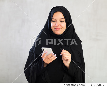 Smiling young woman dressed in traditional islamic clothes looking up something on her phone Smiling young woman dressed in traditional islamic clothes looking up something on her phone 139225077