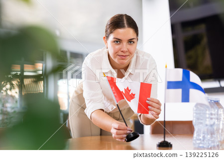 Positive young woman putting flag of Canada on the table with flag of Finland in conference room 139225126