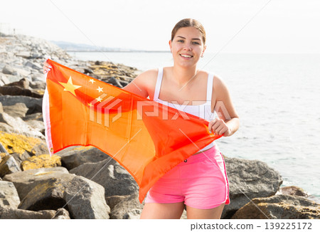 Young woman holding China waving flag at coastline against of bright sea Young woman holding China waving flag at coastline against of bright sea 139225172