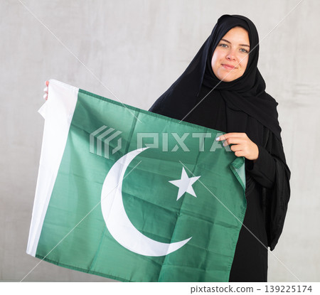 Balanced smiling Muslim woman in traditional black hijab holds flag of Pakistan 139225174