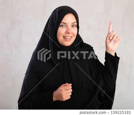 Cheerful young muslim woman dressed in black hijab pointing at side 139225183