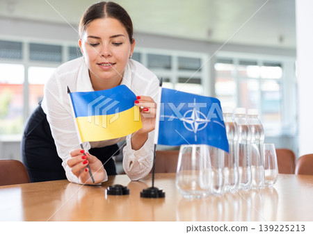 Young woman puts flags of Ukraine and NATO on table in office Young woman puts flags of Ukraine and NATO on table in office 139225213