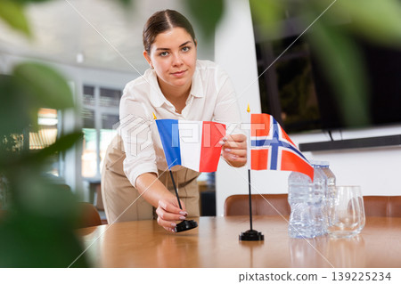 Positive young woman putting flag of France on the table with flag of Norway in conference room 139225234