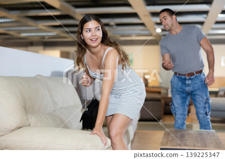 Young woman carefully examines sofa before buying and checks elasticity of surface with hand Young woman carefully examines sofa before buying and checks elasticity of surface with hand 139225347