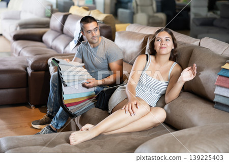 In store wife settled down with legs to test of sofa in use,husband examines upholstery fabric 139225403