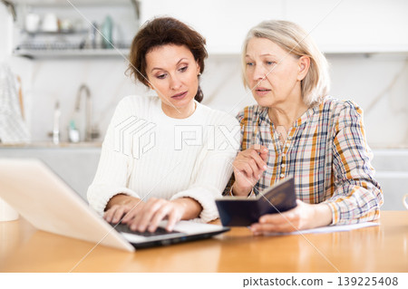 Old woman showing her passport to middle-aged woman sitting next to her and working on laptop 139225408