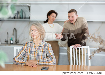 Upset old woman sitting at table with her back to angry middle-aged man and woman Upset old woman sitting at table with her back to angry middle-aged man and woman 139225681