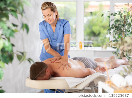 Female masseur performs back stroking and patting during massage session for male client Female masseur performs back stroking and patting during massage session for male client 139225687