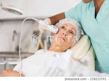 Cosmetologist performing anti-aging face hardware therapy procedure for aged female client in clinic of aesthetic medicine Cosmetologist performing anti-aging face hardware therapy procedure for aged female client in clinic of aesthetic medicine 139225741