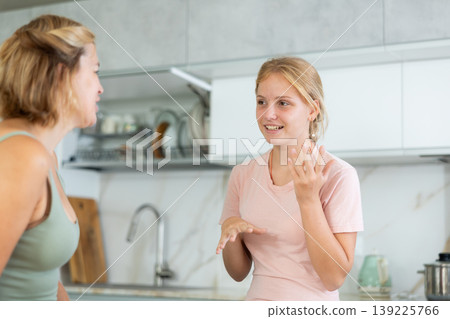 Cheerful teenage girl chatting friendly to mother in home kitchen Cheerful teenage girl chatting friendly to mother in home kitchen 139225766