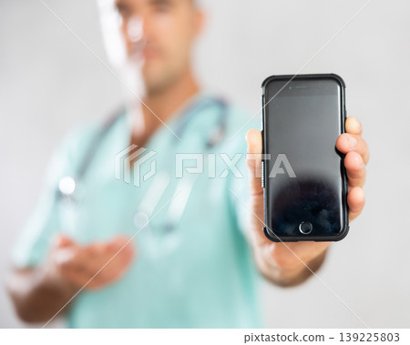 Male doctor in medical uniform posing with mobile phone in medical room Male doctor in medical uniform posing with mobile phone in medical room 139225803