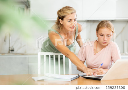 Mother looks at laptop and prompts and helps teen girl to write in notebook to complete task Mother looks at laptop and prompts and helps teen girl to write in notebook to complete task 139225808