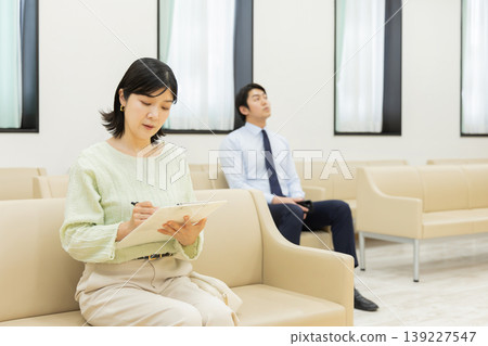 一名女子在候診室填寫醫療問卷,一名商人正在等待輪到自己。 一名女子在候診室填寫醫療問卷,一名商人正在等待輪到自己。 139227547