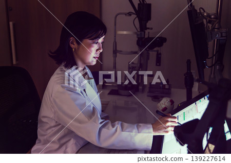A female ophthalmologist operates an electronic medical record system in a dimly lit examination room (the monitor is a test screen). 139227614
