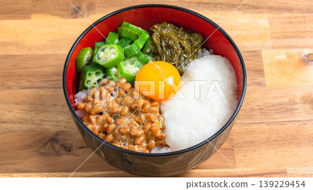 Sticky Rice Bowl: Rice topped with natto, okra, grated yam, mekabu seaweed, and egg yolk, against a wood grain background. Sticky Rice Bowl: Rice topped with natto, okra, grated yam, mekabu seaweed, and egg yolk, against a wood grain background. 139229454