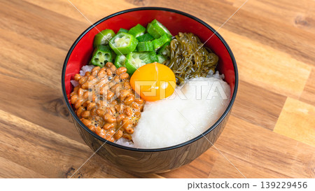 Sticky Rice Bowl: Rice topped with natto, okra, grated yam, mekabu seaweed, and egg yolk, against a wood grain background. Sticky Rice Bowl: Rice topped with natto, okra, grated yam, mekabu seaweed, and egg yolk, against a wood grain background. 139229456