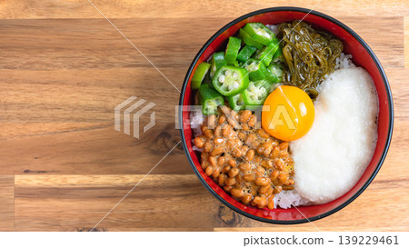糯米飯碗：米飯上放有納豆、秋葵、磨碎的山藥、海苔和蛋黃，背景是木紋。 139229461