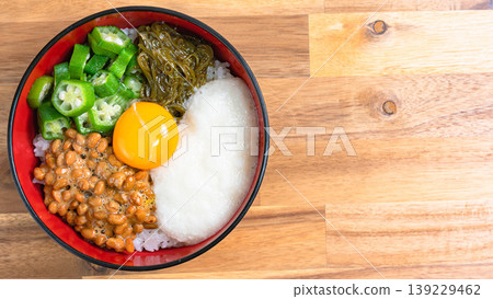 Sticky Rice Bowl: Rice topped with natto, okra, grated yam, mekabu seaweed, and egg yolk, against a wood grain background. Sticky Rice Bowl: Rice topped with natto, okra, grated yam, mekabu seaweed, and egg yolk, against a wood grain background. 139229462
