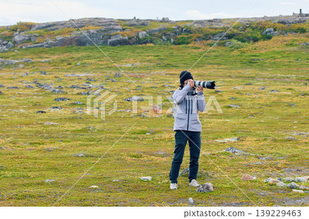 A photographer girl with a professional photo camera walks through a rocky area and on the green meadow in cloudy weather 139229463