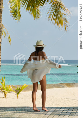A beautiful tanned girl is wearing a hat and swimsuit with an athletic physique stands on the beach of the villa on a sunny day in the Maldives, she is happy and posing 139229581