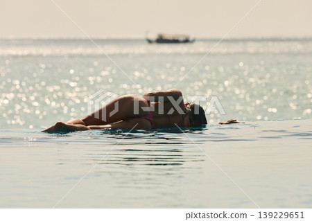 A beautiful tanned girl with an athletic physique in a swimsuit is at the edge of the pool of the villa pool on a sunset in the Maldives, she is happy and posing A beautiful tanned girl with an athletic physique in a swimsuit is at the edge of the pool of the villa pool on a sunset in the Maldives, she is happy and posing 139229651