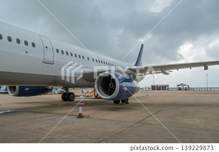 White commercial jetliner parked on airport apron with blue engine against grey clouds 139229873