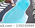 Top view of a curved turquoise swimming pool surrounded by loungers, chairs, and a shaded umbrella on a tiled patio, evoking a calm resort atmosphere and relaxation.  139230999