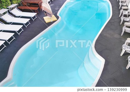 Top view of a curved turquoise swimming pool surrounded by loungers, chairs, and a shaded umbrella on a tiled patio, evoking a calm resort atmosphere and relaxation. Top view of a curved turquoise swimming pool surrounded by loungers, chairs, and a shaded umbrella on a tiled patio, evoking a calm resort atmosphere and relaxation. 139230999