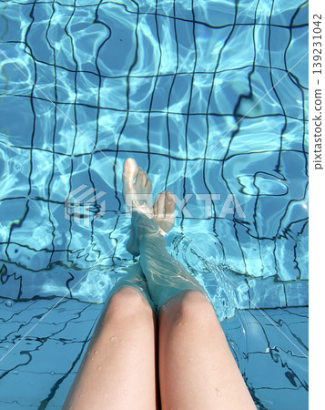 In the crystal clear, sunlit, sparkling blue water of the pool, the woman comfortably crossed her bare legs and feet. In the crystal clear, sunlit, sparkling blue water of the pool, the woman comfortably crossed her bare legs and feet. 139231042
