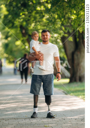 Conception of love and care, standing. Father that is amputee with prosthesis is with his little son in the park Conception of love and care, standing. Father that is amputee with prosthesis is with his little son in the park 139231123