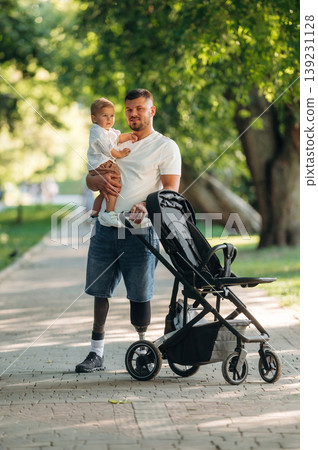 With a cradle. Father that is amputee with prosthesis is with his little son in the park 139231128