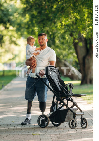 With a cradle. Father that is amputee with prosthesis is with his little son in the park With a cradle. Father that is amputee with prosthesis is with his little son in the park 139231129