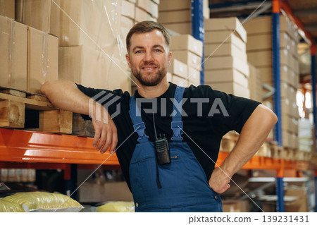 Leaning by the shelve, standing and posing. Man in uniform is working in the storage with packages for shipment Leaning by the shelve, standing and posing. Man in uniform is working in the storage with packages for shipment 139231431