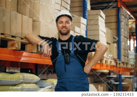 Leaning by the shelve, standing and posing. Man in uniform is working in the storage with packages for shipment Leaning by the shelve, standing and posing. Man in uniform is working in the storage with packages for shipment 139231433