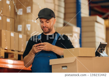 Time for a break, using smartphone. Man in uniform is working in the storage with packages for shipment Time for a break, using smartphone. Man in uniform is working in the storage with packages for shipment 139231442