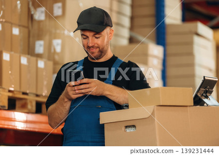Texting by phone. Man in uniform is working in the storage with packages for shipment Texting by phone. Man in uniform is working in the storage with packages for shipment 139231444