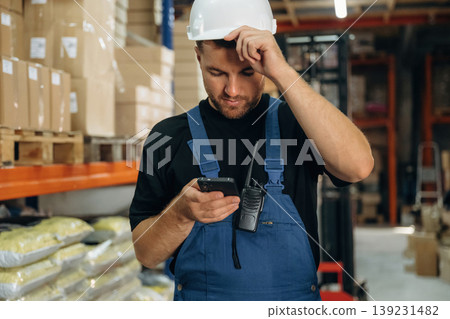 Uses smartphone, holding it in hands. Man in uniform is working in the storage with packages for shipment Uses smartphone, holding it in hands. Man in uniform is working in the storage with packages for shipment 139231482