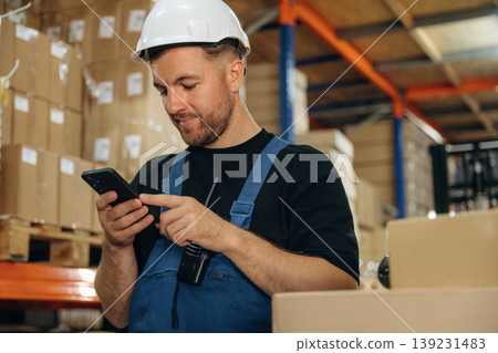 Uses smartphone, holding it in hands. Man in uniform is working in the storage with packages for shipment Uses smartphone, holding it in hands. Man in uniform is working in the storage with packages for shipment 139231483