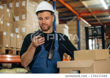Uses smartphone, holding it in hands. Man in uniform is working in the storage with packages for shipment 139231484