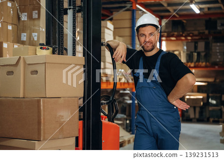Model in hard hat is leaning on the forklift and smiling. Man in uniform is working in the storage with packages for shipment Model in hard hat is leaning on the forklift and smiling. Man in uniform is working in the storage with packages for shipment 139231513