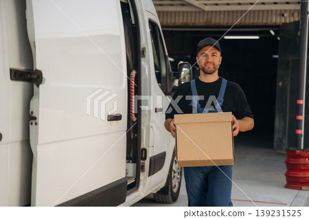 Looking forward, standing and holding the box. Delivery man is by the bus near garage outdoors Looking forward, standing and holding the box. Delivery man is by the bus near garage outdoors 139231525