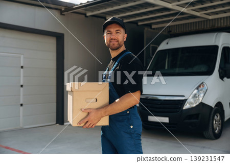 In black cap, with box in hands. Delivery man is by the bus near garage outdoors 139231547
