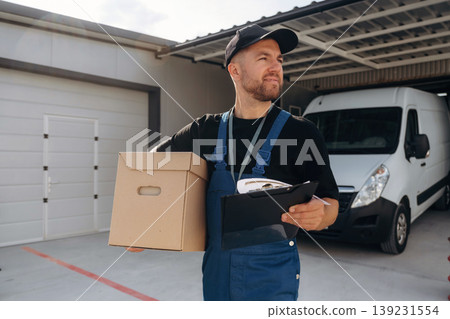 In black cap, with box in hands. Delivery man is by the bus near garage outdoors In black cap, with box in hands. Delivery man is by the bus near garage outdoors 139231554