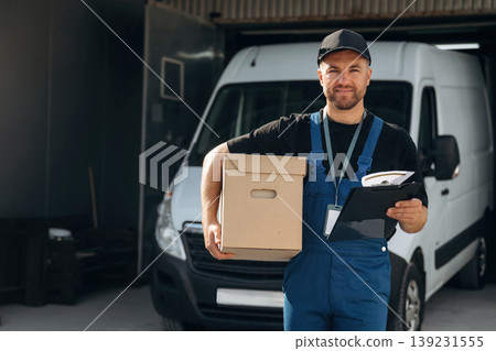 In black cap, with box in hands. Delivery man is by the bus near garage outdoors In black cap, with box in hands. Delivery man is by the bus near garage outdoors 139231555
