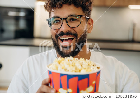 Delicious popcorn in a bucket. Man is in the modern domestic room at home Delicious popcorn in a bucket. Man is in the modern domestic room at home 139231850