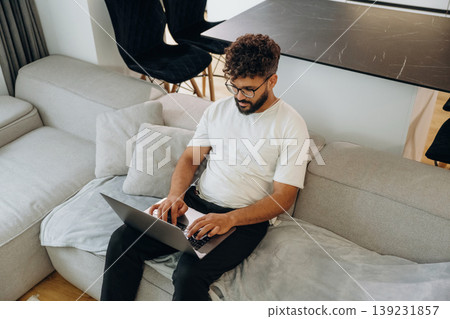 Laptop is on the knees, guy is sitting on the sofa. Man is in the modern domestic room at home 139231857