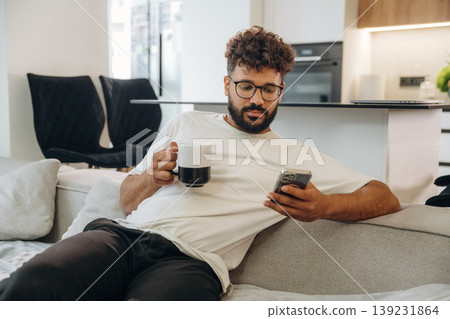 Cup of drink and smartphone are in hands. Man is in the modern domestic room at home Cup of drink and smartphone are in hands. Man is in the modern domestic room at home 139231864