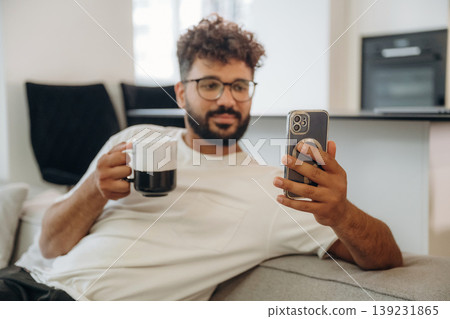 Reading social media, using phone, holding cup of drink. Man is in the modern domestic room at home Reading social media, using phone, holding cup of drink. Man is in the modern domestic room at home 139231865